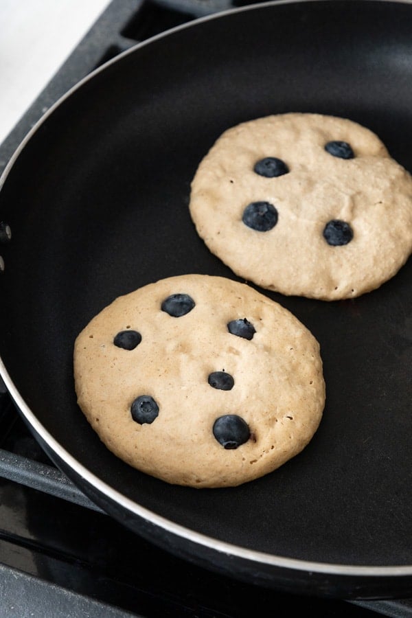 two vegan blueberry banana pancakes frying on a skillet