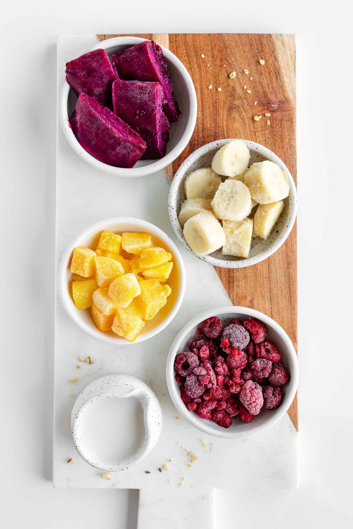 marble serving board topped with bowls of dragon fruit, frozen bananas, frozen mango chunks, frozen raspberries, and coconut milk