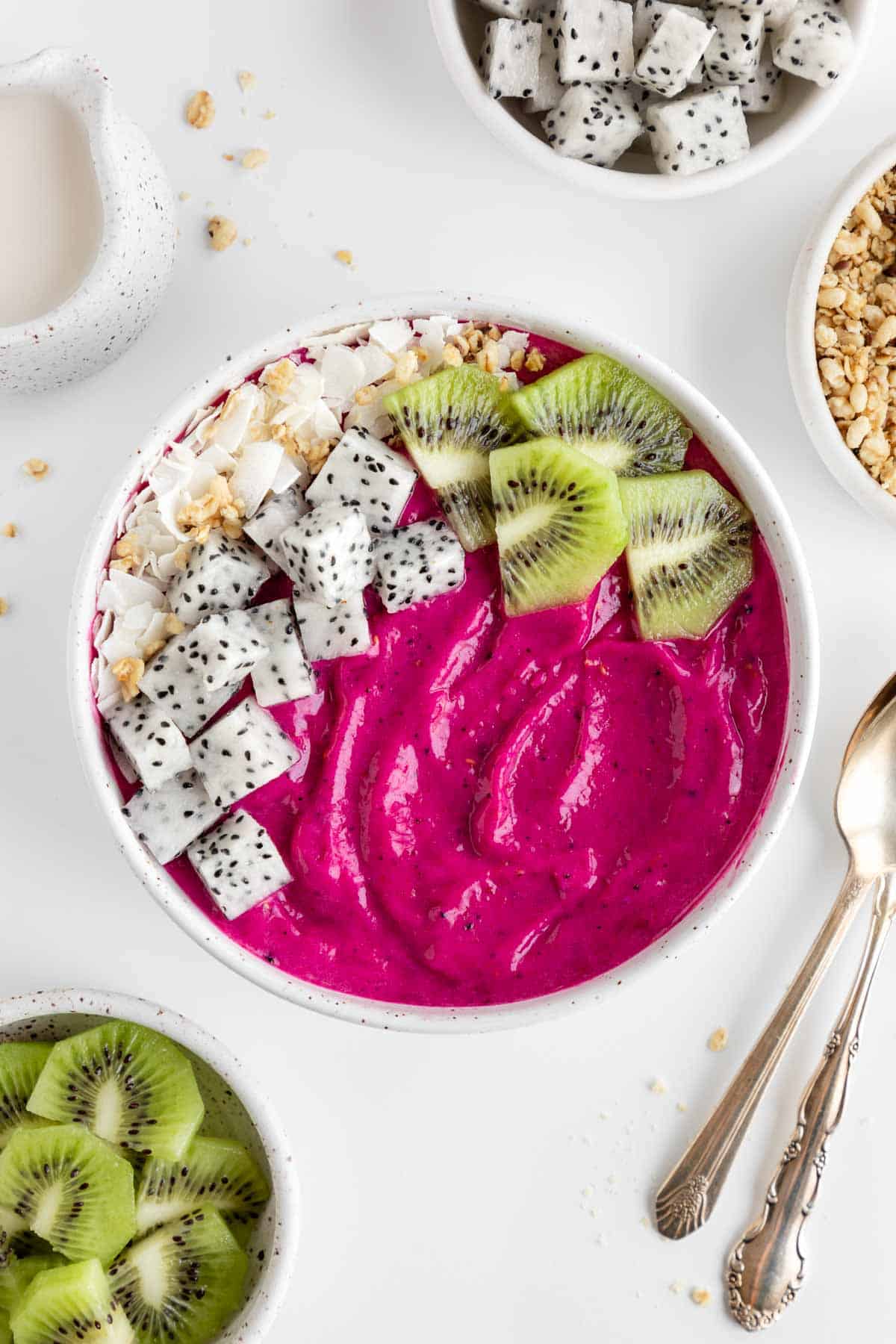 a dragon fruit smoothie bowl beside vintage spoons and bowls of fresh fruit