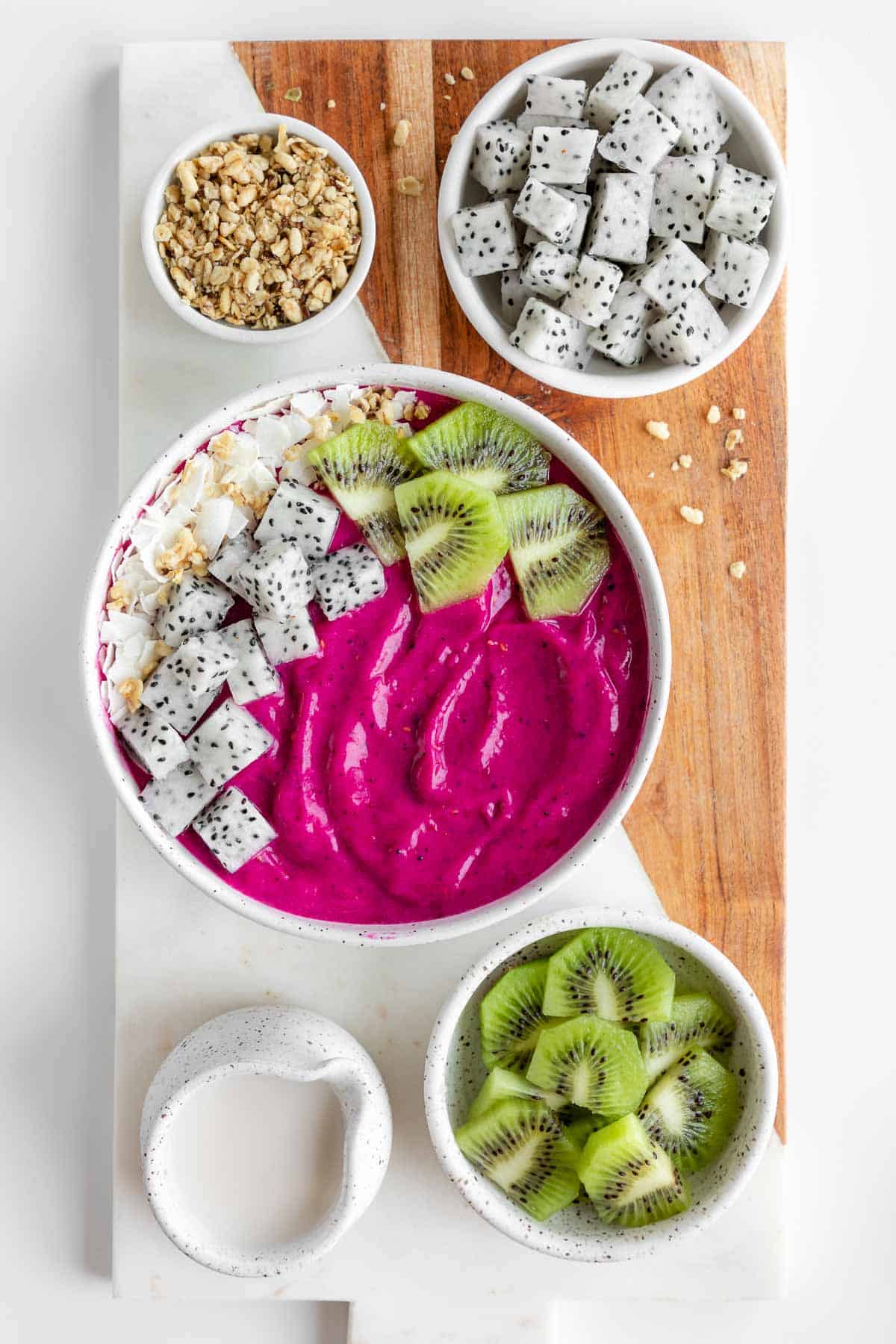 a pitaya bowl on a marble and wood serving board with dragon fruit, kiwi, granola, and coconut milk
