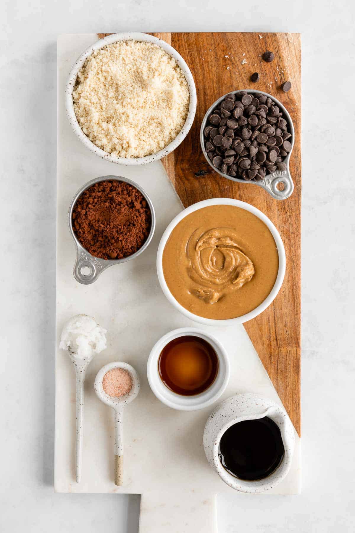 a marble serving board topped with bowls of almond flour, peanut butter, cocoa powder, maple syrup, chocolate chips, and vanilla extract