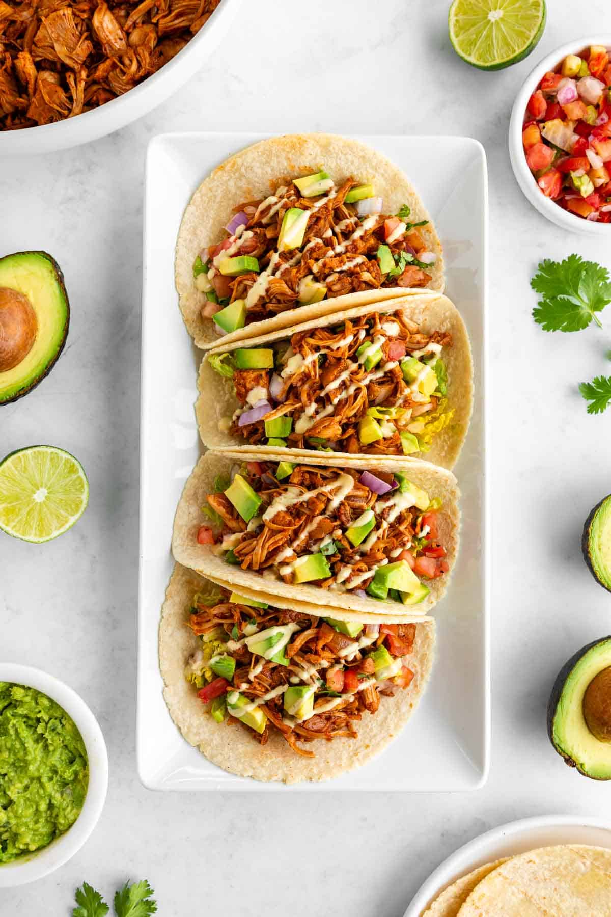 four vegan jackfruit tacos on a white rectangular plate surrounded by ingredients