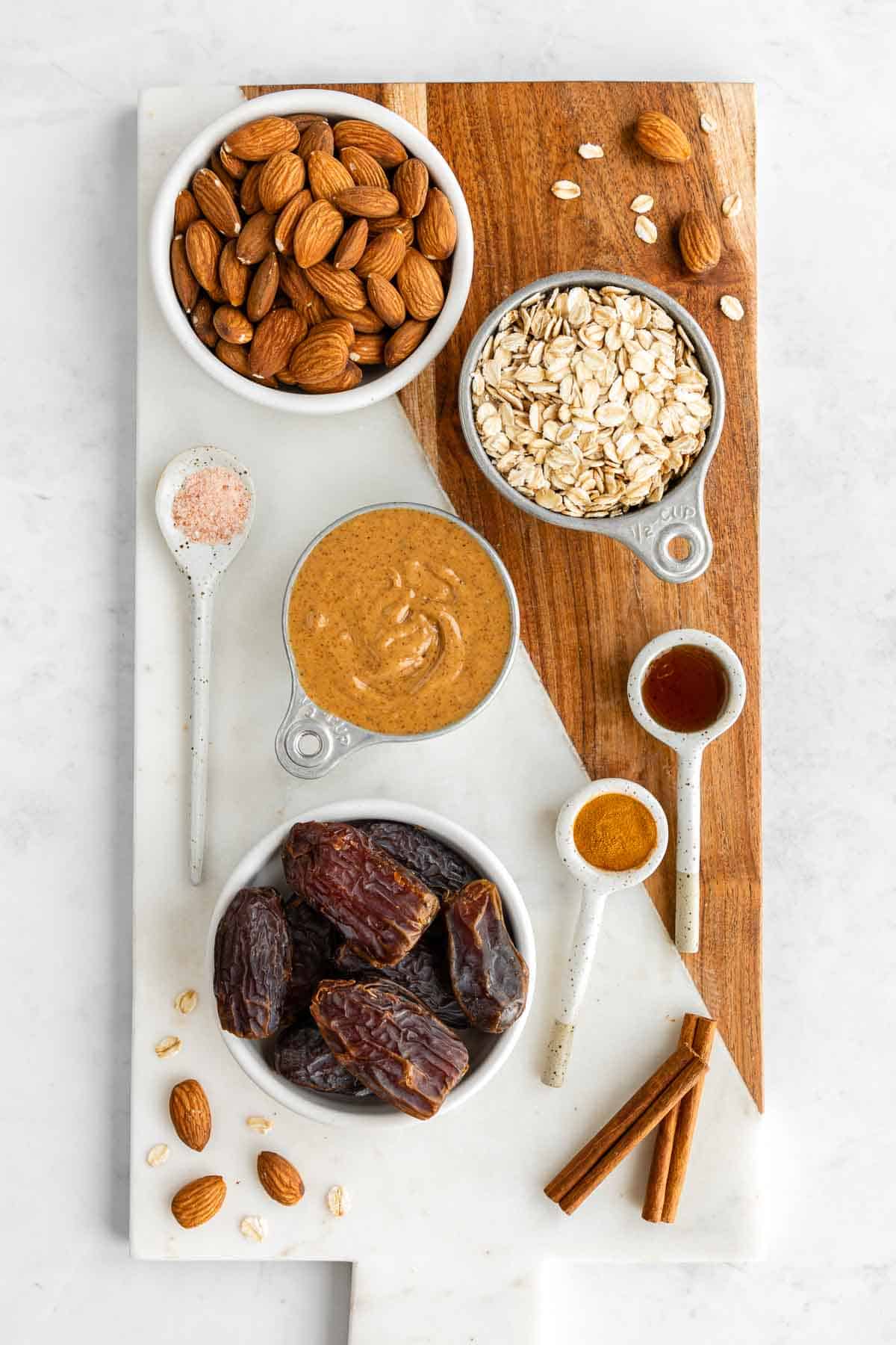 a marble and wood board topped with bowls of almonds, oats, dates, vanilla, cinnamon, and salt