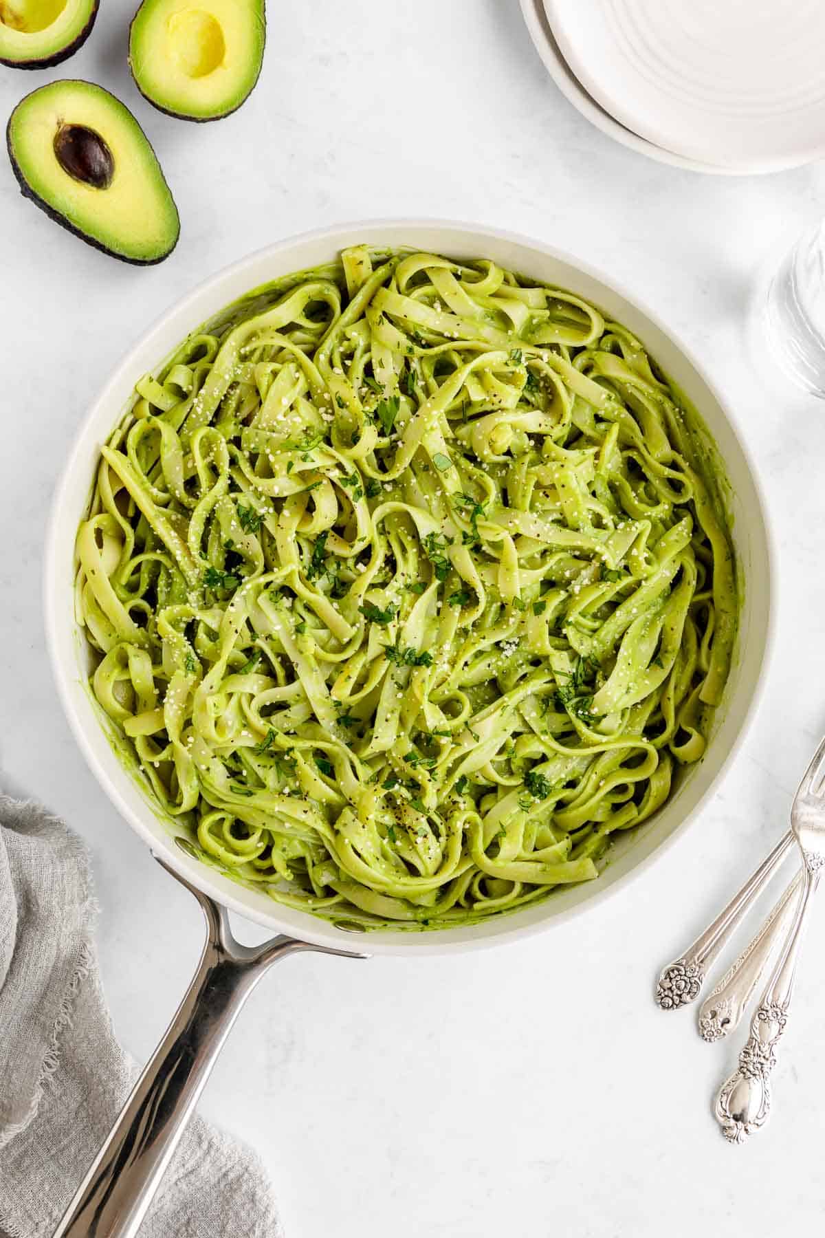 avocado pasta in a pot beside white plates, sliced avocados, and silverware