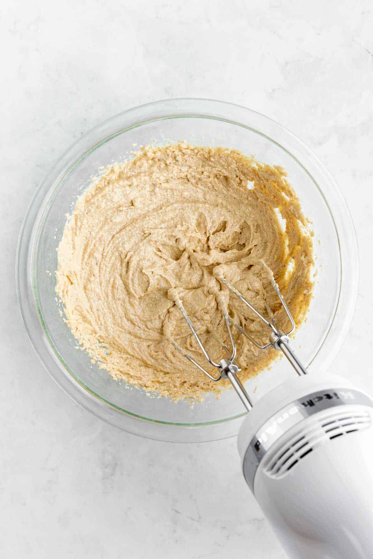 a handheld electric mixing beating vegan butter and sugar together in a glass bowl