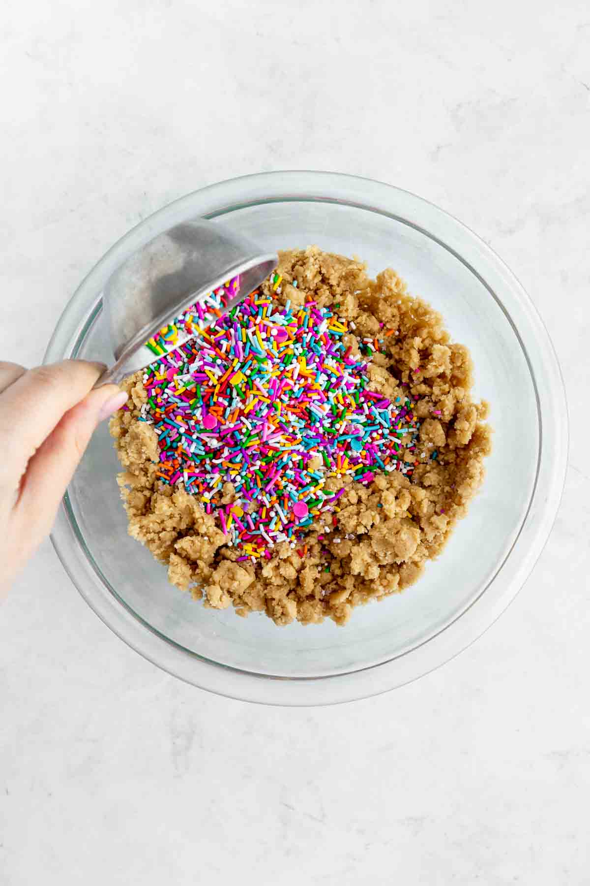pouring vegan rainbow sprinkles over a bowl of edible cookie dough in a glass bowl