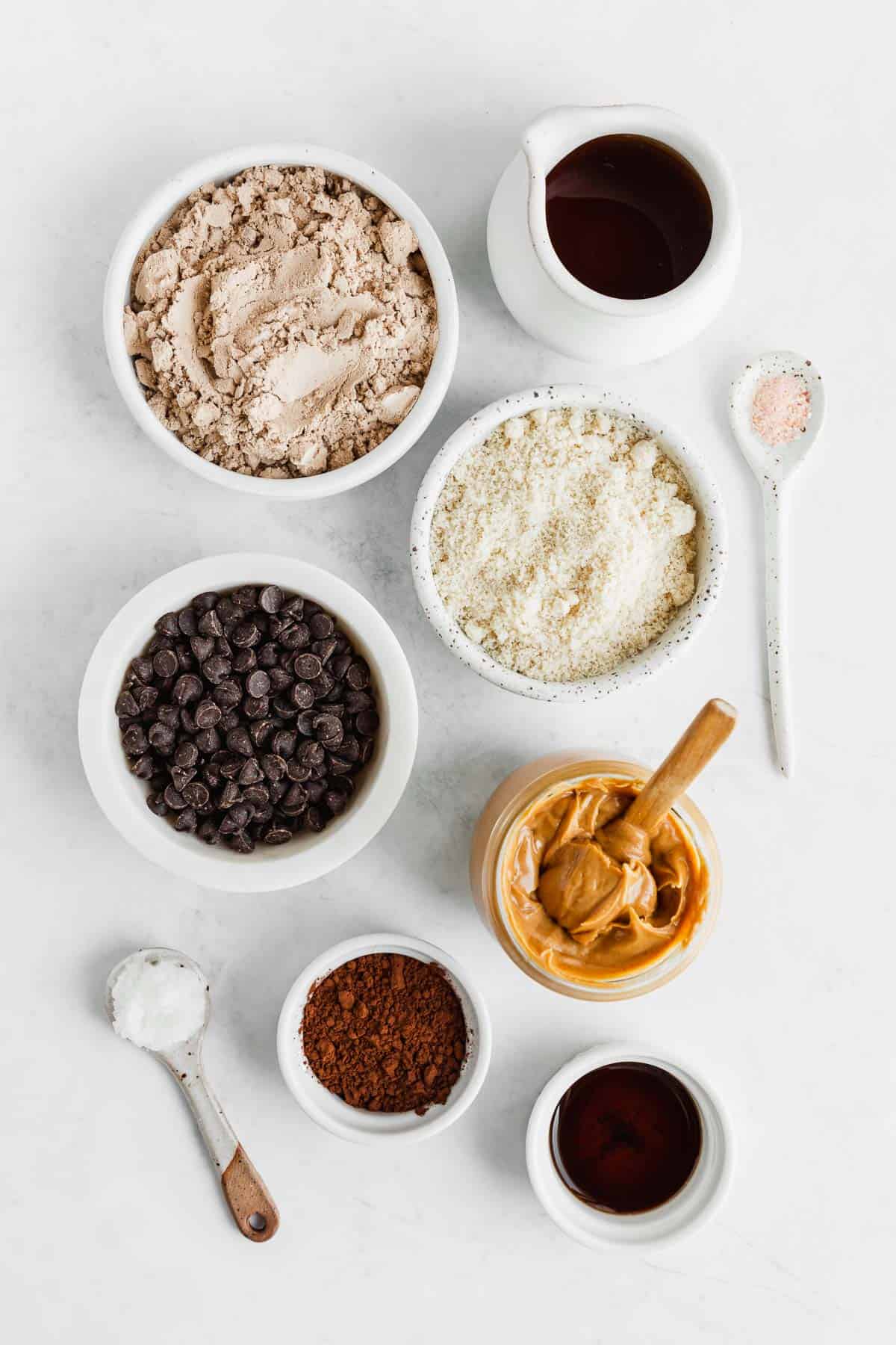 bowls of chocolate protein powder, cocoa powder, almond flour, peanut butter, chocolate chips, maple syrup, and vanilla extract