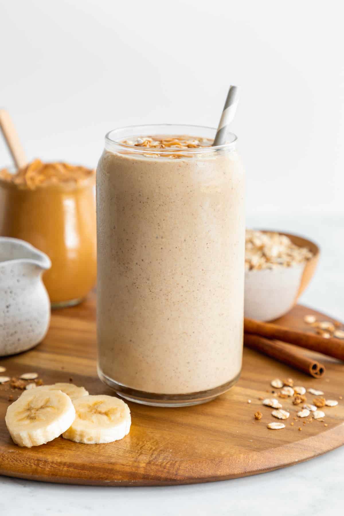 an oatmeal smoothie inside a glass surrounded by sliced banana, a jar of peanut butter, oats, and cinnamon sticks