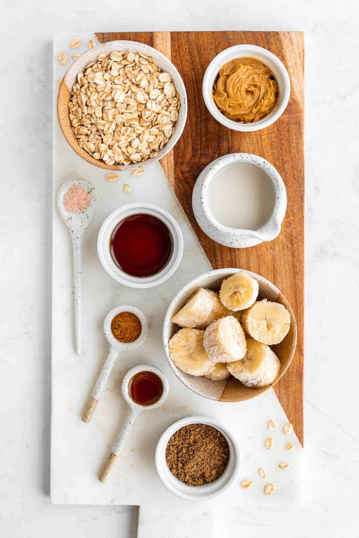 bowls of rolled oats, nut butter, almond milk, chopped frozen banana, cinnamon, ground flaxseed, vanilla extract, and maple syrup on a marble and wood board