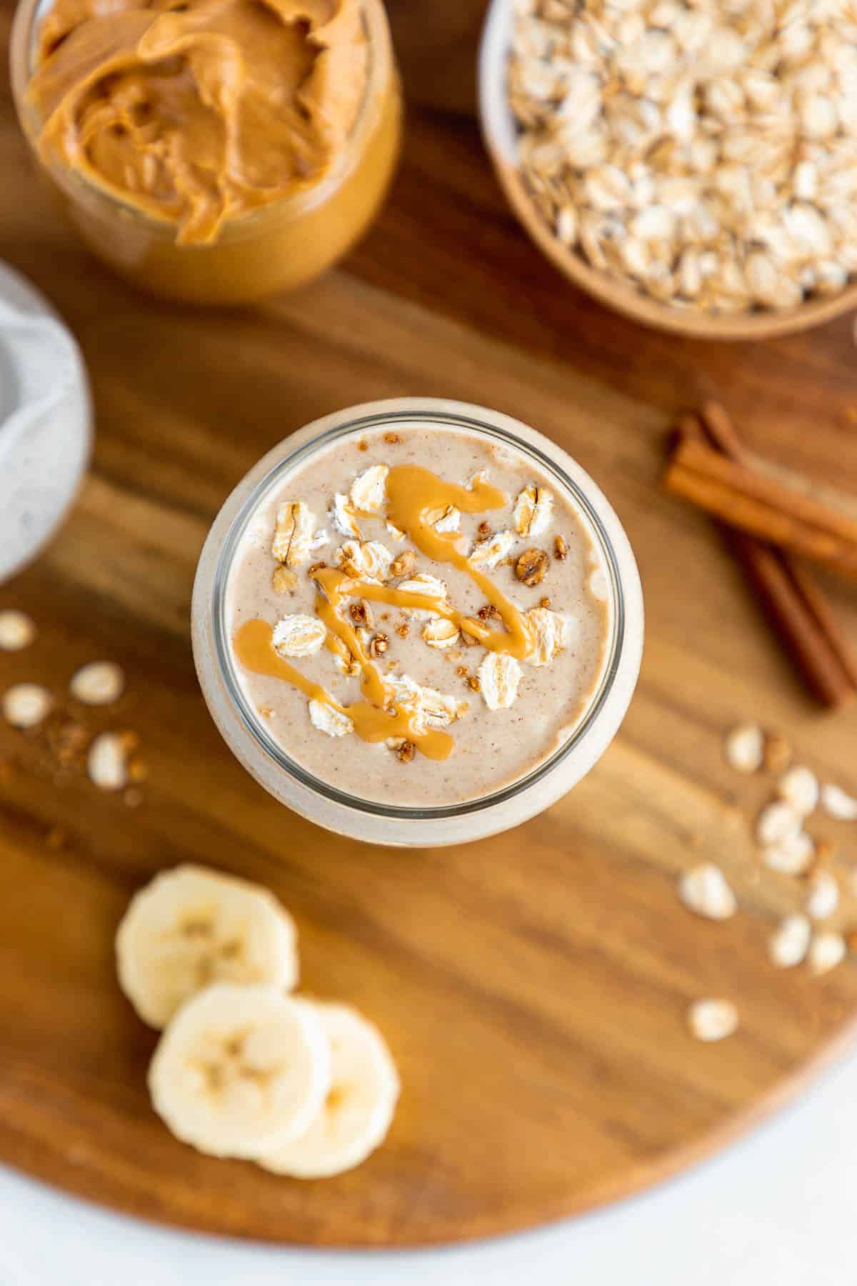 overhead photo of a peanut butter oatmeal smoothie inside a glass, surrounded by sliced banana, rolled oats, and oat milk