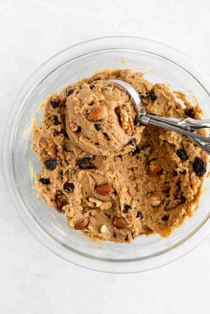 a metal scooper scooping trail mix cookie dough in a glass bowl
