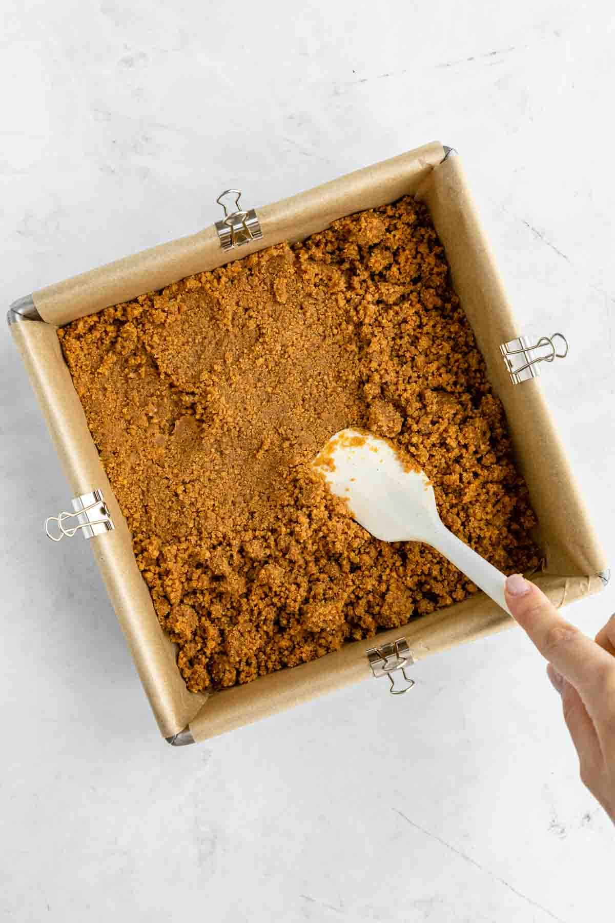 a spatula pressing graham cracker crust into a square baking dish