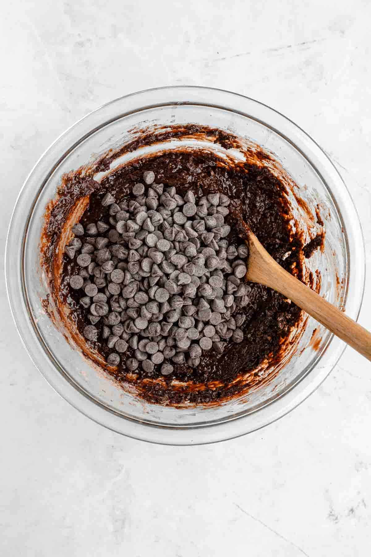 a wooden spoon mixing chocolate chips into vegan brownie batter inside a glass bowl