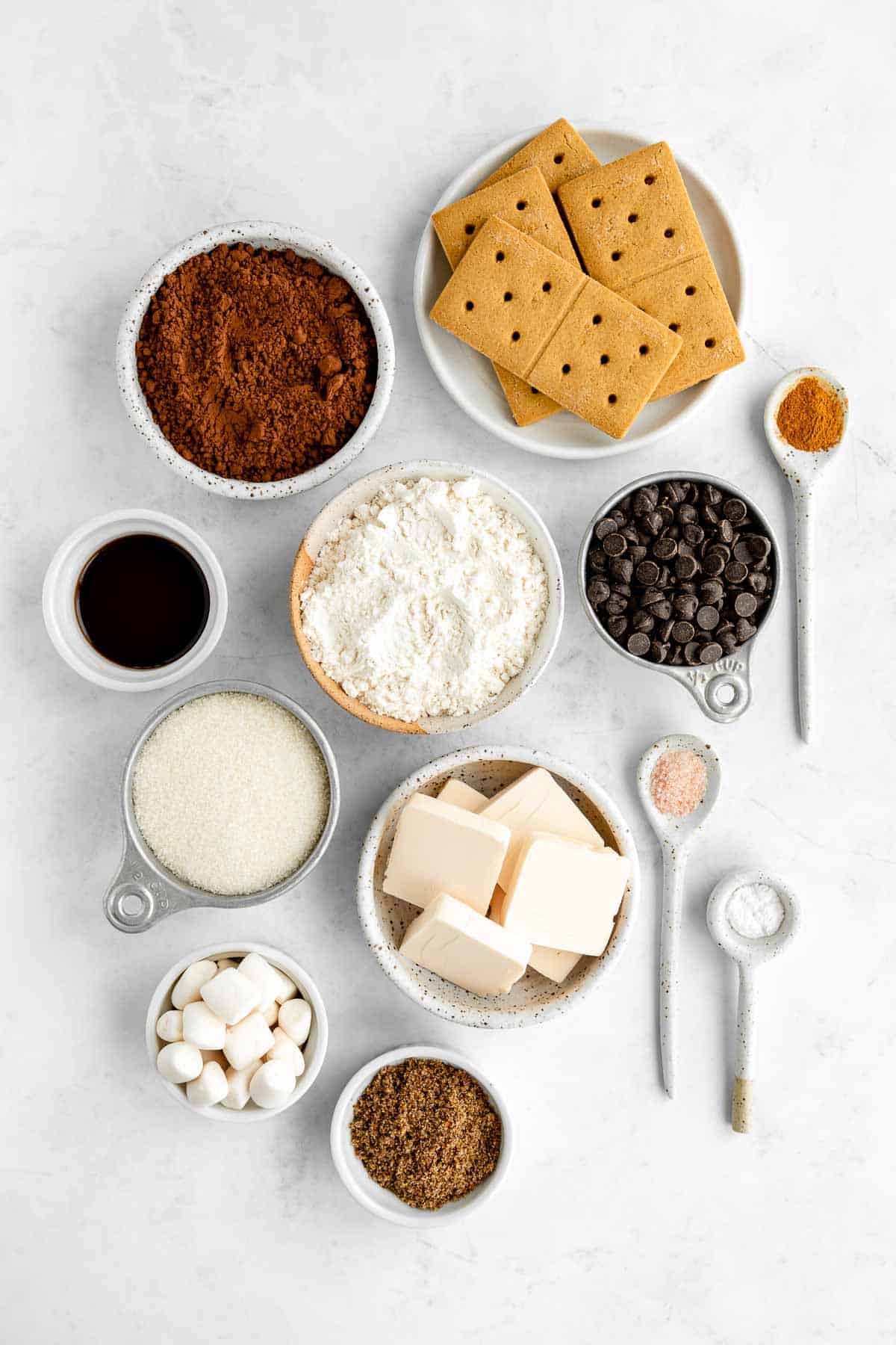 bowls filled with graham crackers, vegan marshmallows, chocolate chips, dairy-free butter, flour, sugar, cocoa powder, and vanilla