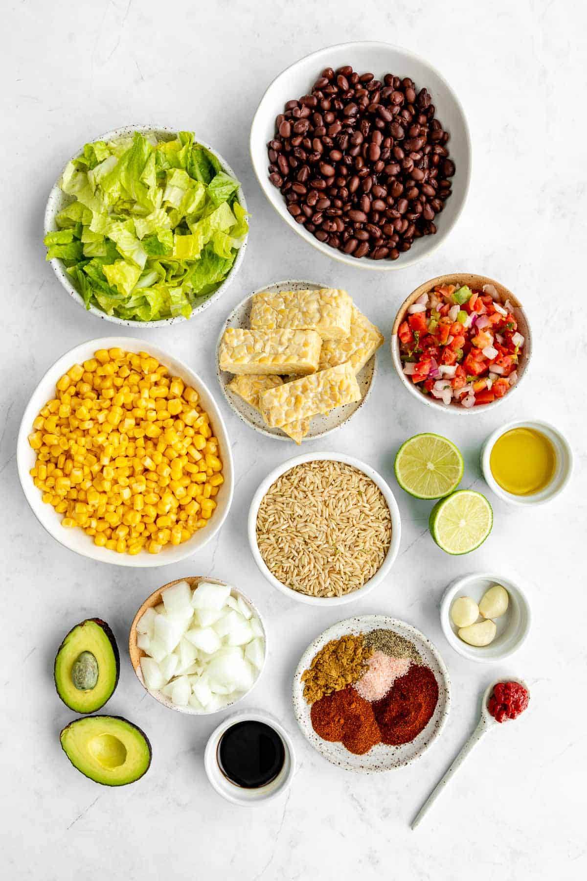 bowls of ingredients for a vegan tempeh burrito bowl, including corn, lettuce, black beans, pico de gallo, onion, avocado, and spices