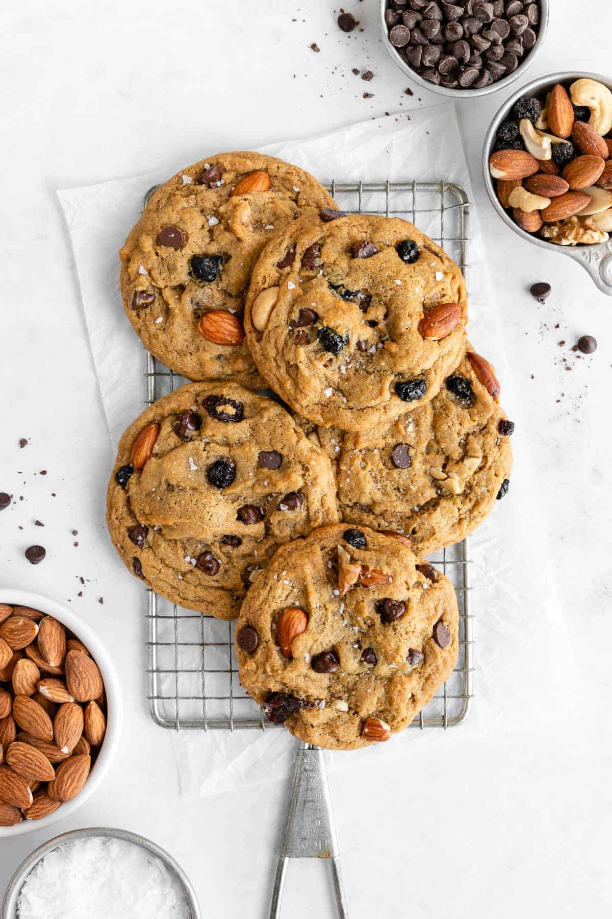 a pile of vegan trail mix cookies on a cooling rack surrounded by bowls of chocolate chips, nuts, and dried fruit