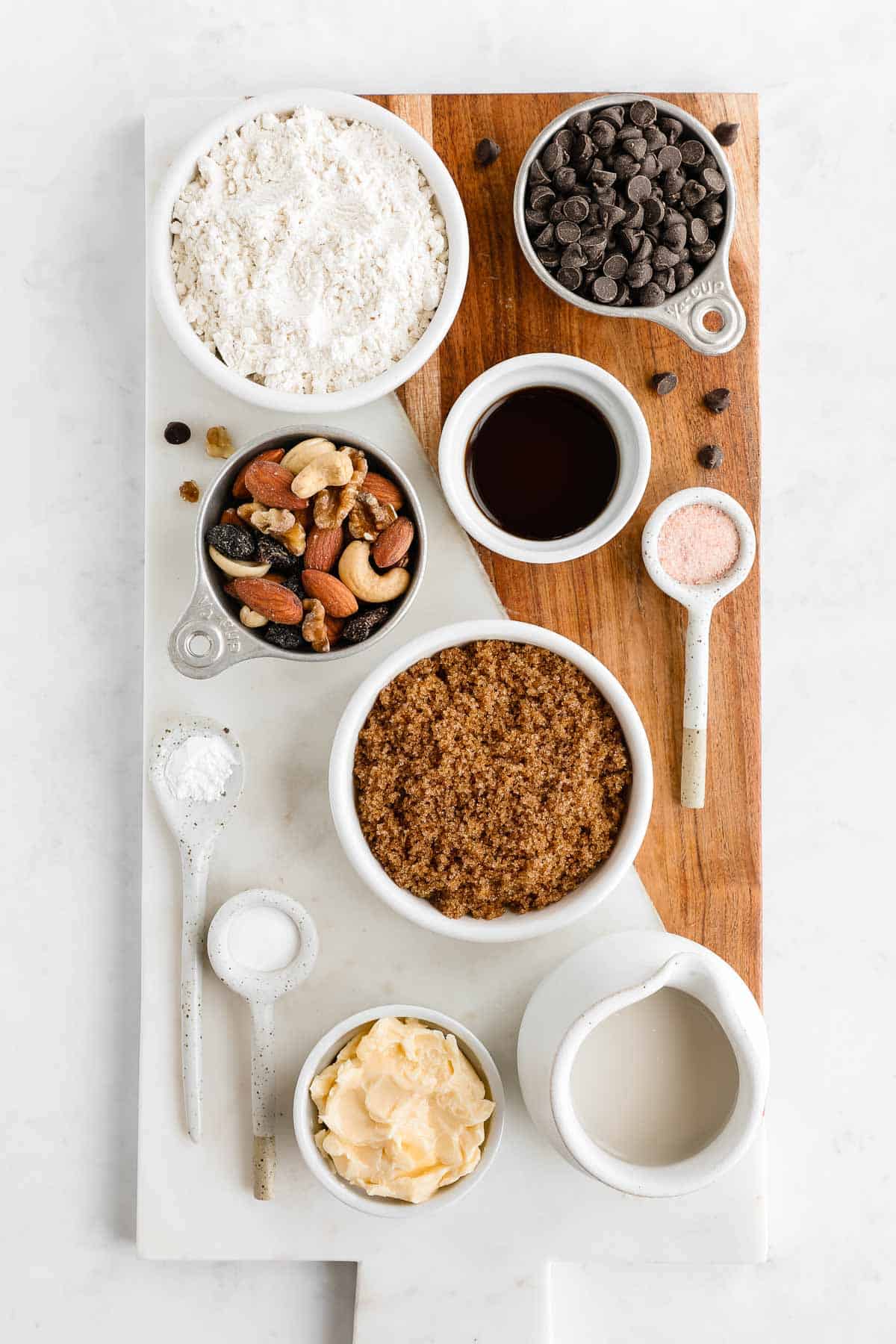 bowls of flour, trail mix, chocolate chips, brown sugar, vegan butter, almond milk, and vanilla extract on a serving board