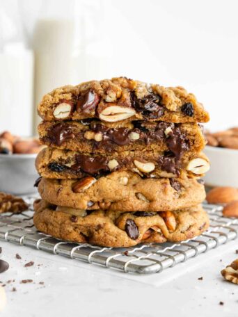 a stack of vegan trail mix cookies sliced in half on a cooling rack surrounded by chocolate chips, mixed nuts, and fruit