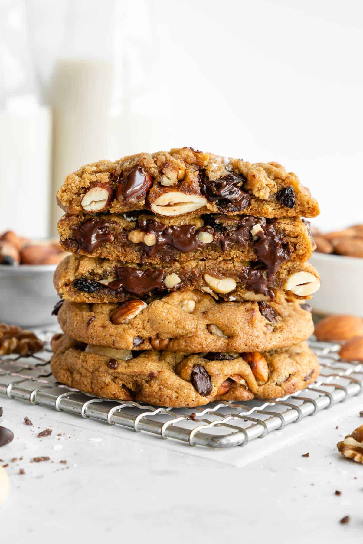 a stack of vegan trail mix cookies sliced in half on a cooling rack surrounded by chocolate chips, mixed nuts, and fruit