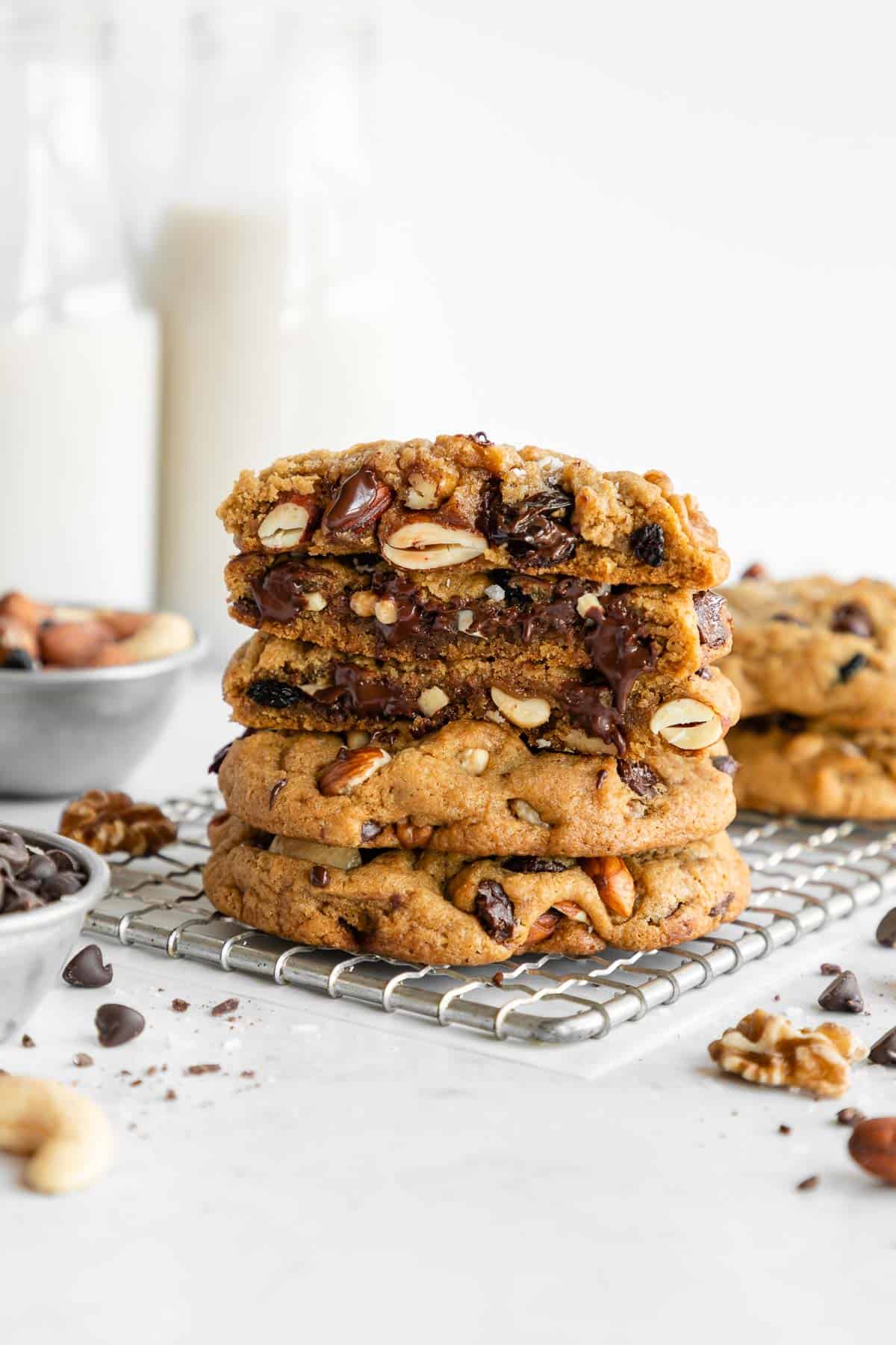 a stack of vegan trail mix cookies sliced in half on a cooling rack surrounded by chocolate chips, mixed nuts, and fruit