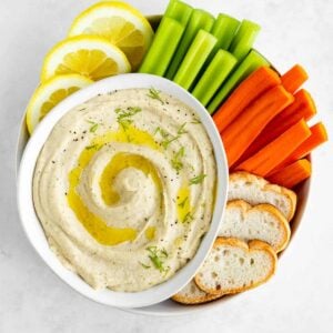white bean dip inside a bowl surrounded by celery, carrots, bread, and lemon