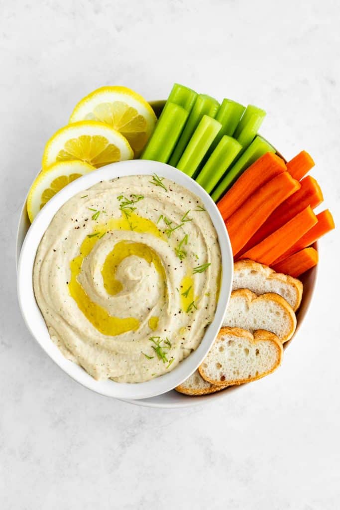 white bean dip inside a bowl surrounded by celery, carrots, bread, and lemon