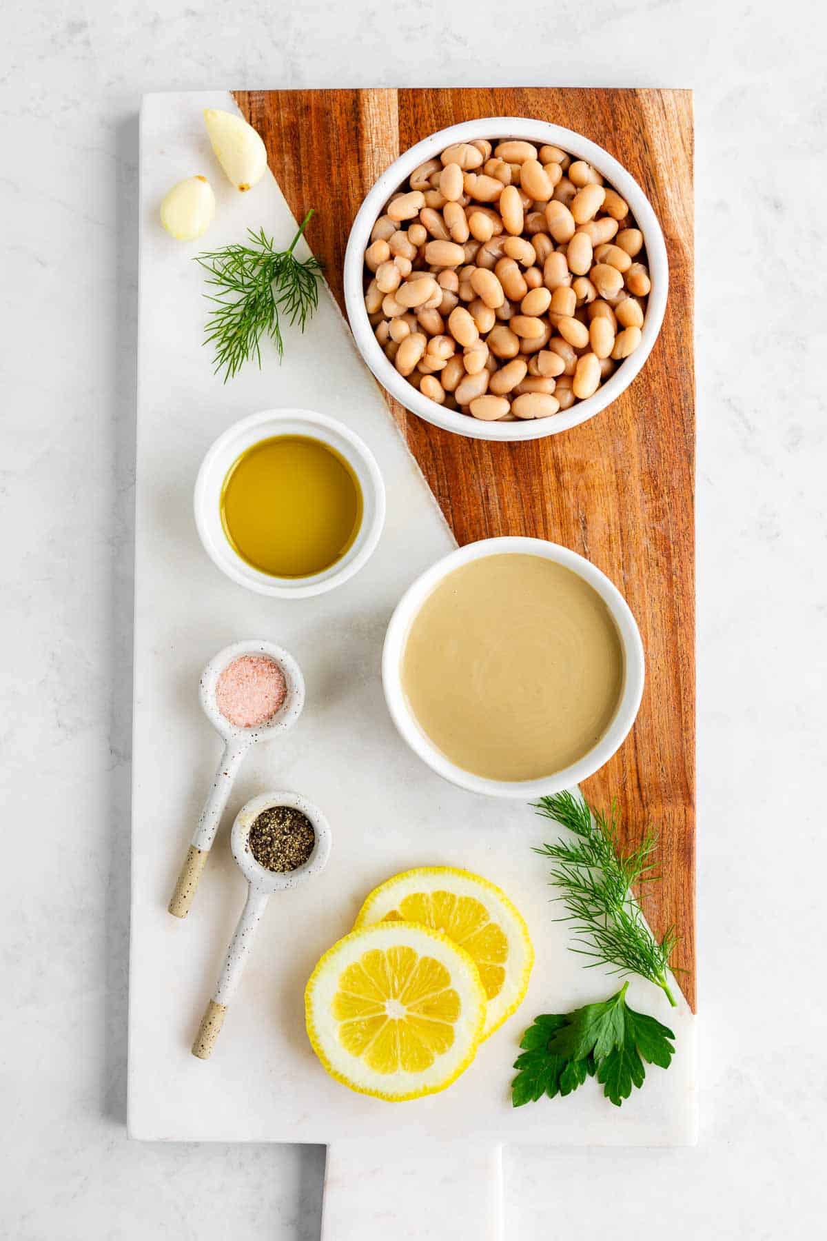 a marble serving board topped with bowls of white beans, tahini, olive oil, a sliced lemon, dill, parsley, and garlic