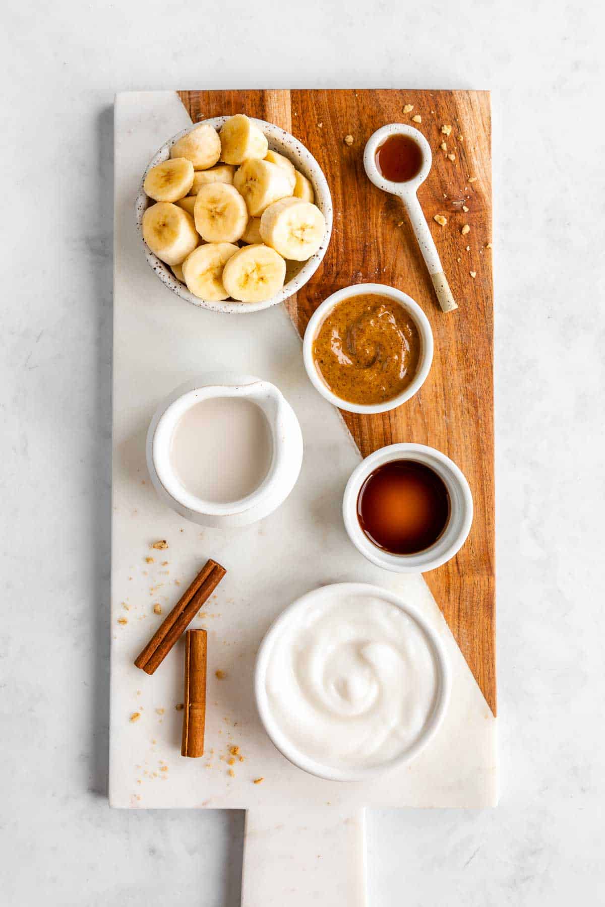 a marble serving board topped with sliced banana, almond butter, almond milk, dairy free yogurt, cinnamon sticks, maple syrup, and vanilla