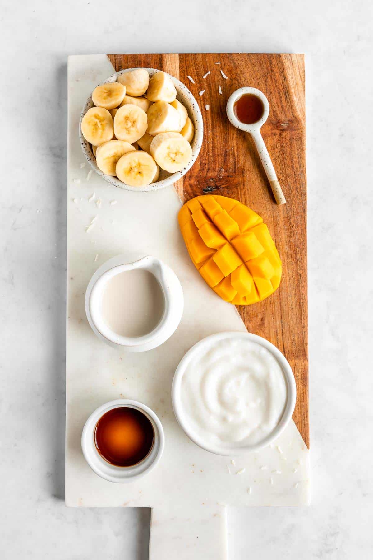 a marble serving board topped with sliced banana, mango, coconut milk, non dairy yogurt, maple syrup, and vanilla extract