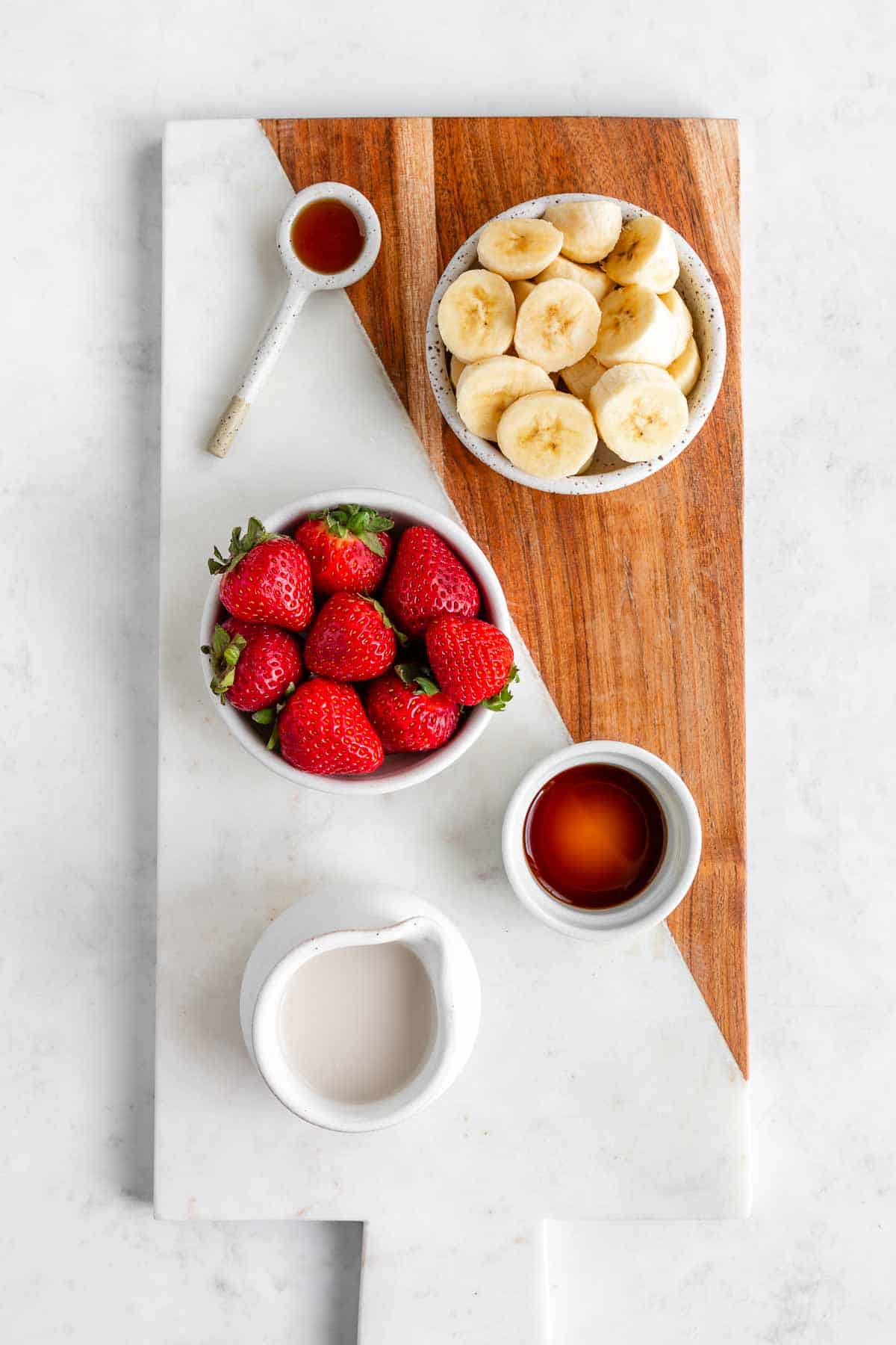 bowls filled with strawberries, banana, vanilla extract, almond milk, and maple syrup on a marble board