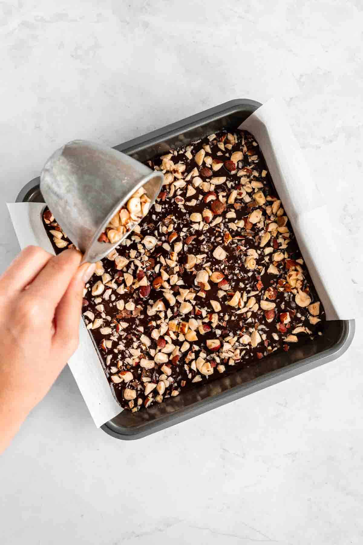 pouring chopped hazelnuts over raw brownies in a square dish