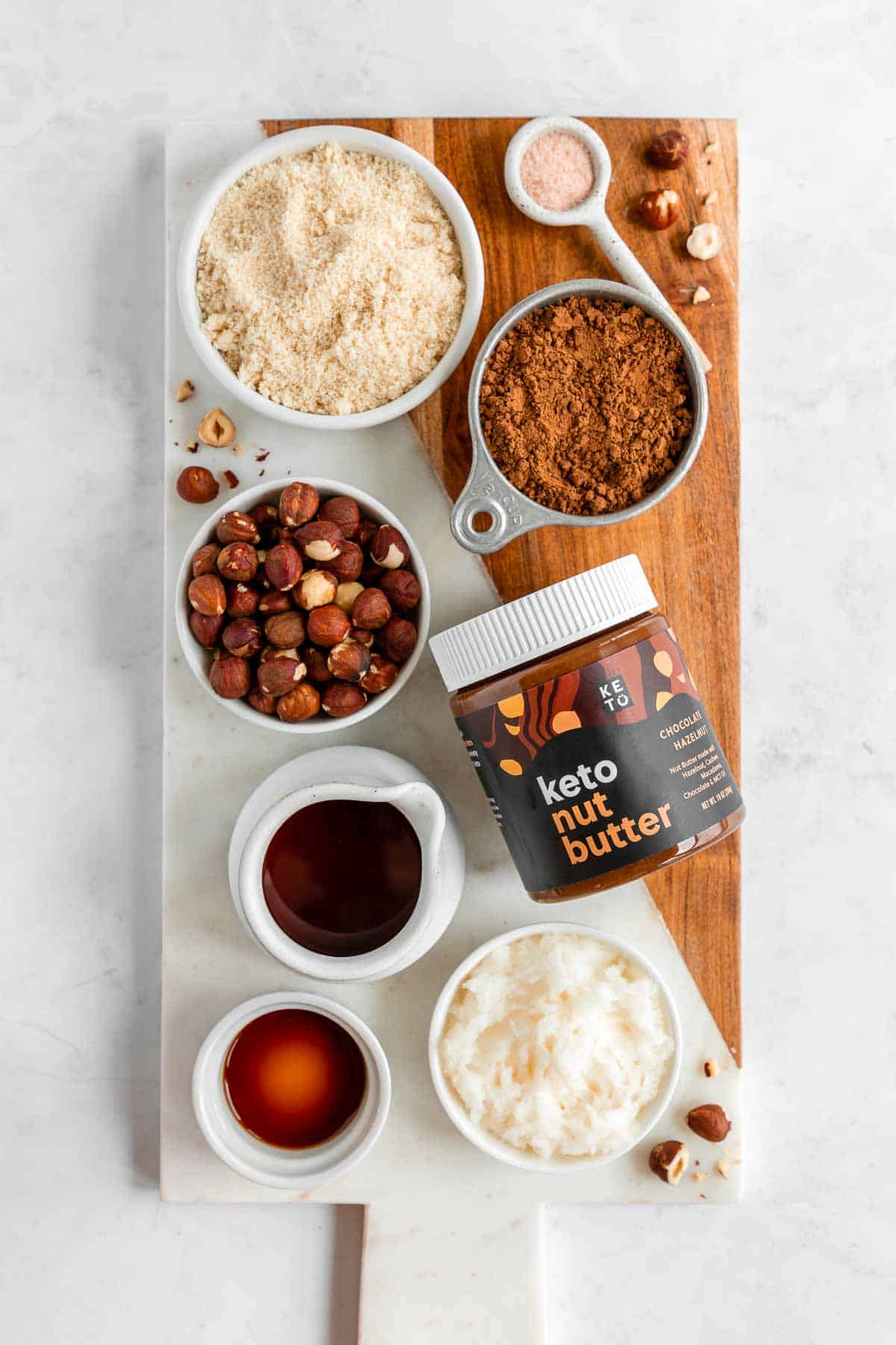 a marble and wood serving board topped with bowls of almond flour, perfect keto hazelnut butter, cacao powder, maple syrup, and hazelnuts