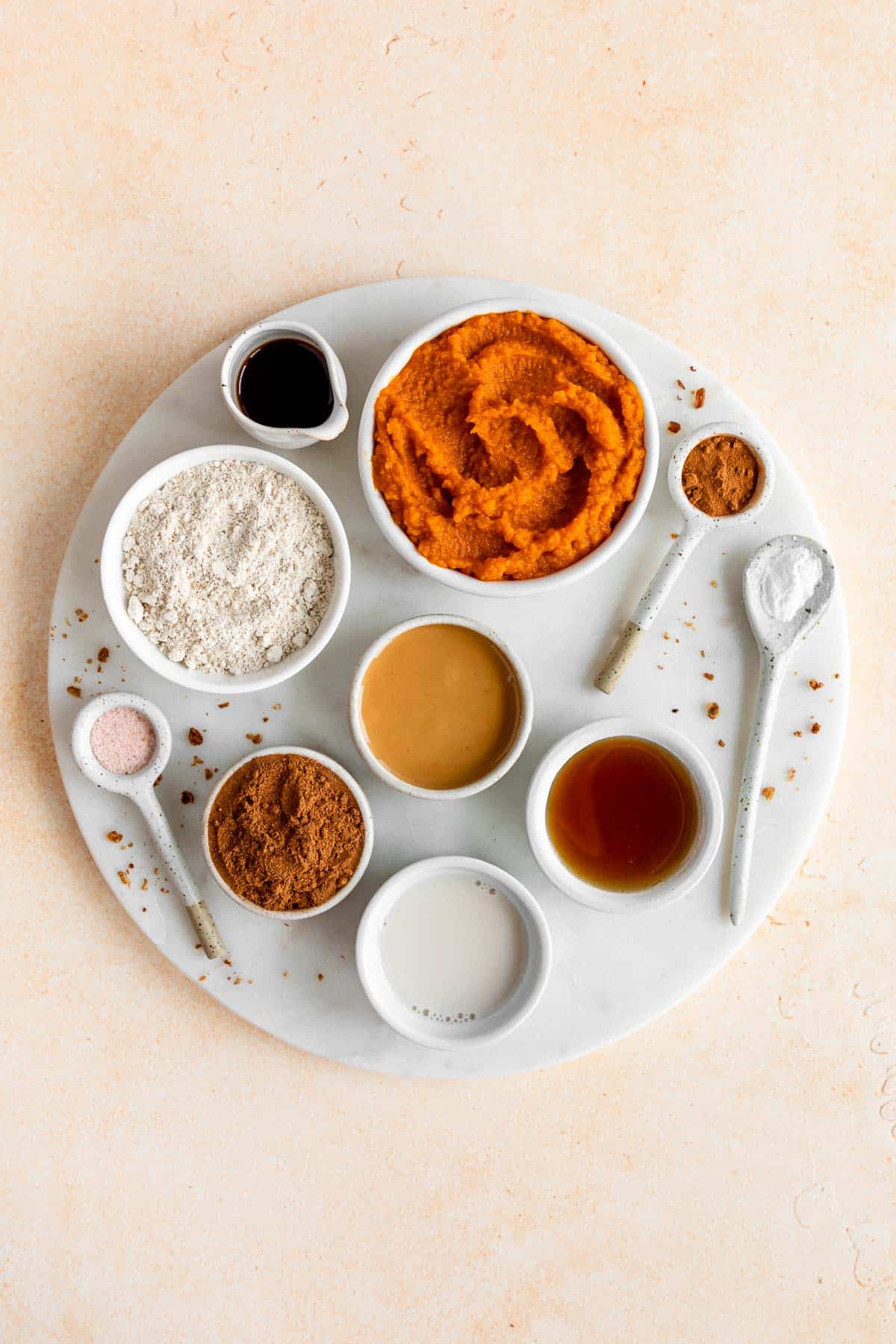 a round marble board with bowls of pumpkin puree, oat flour, almond butter, almond milk, maple syrup, cinnamon, and vanilla