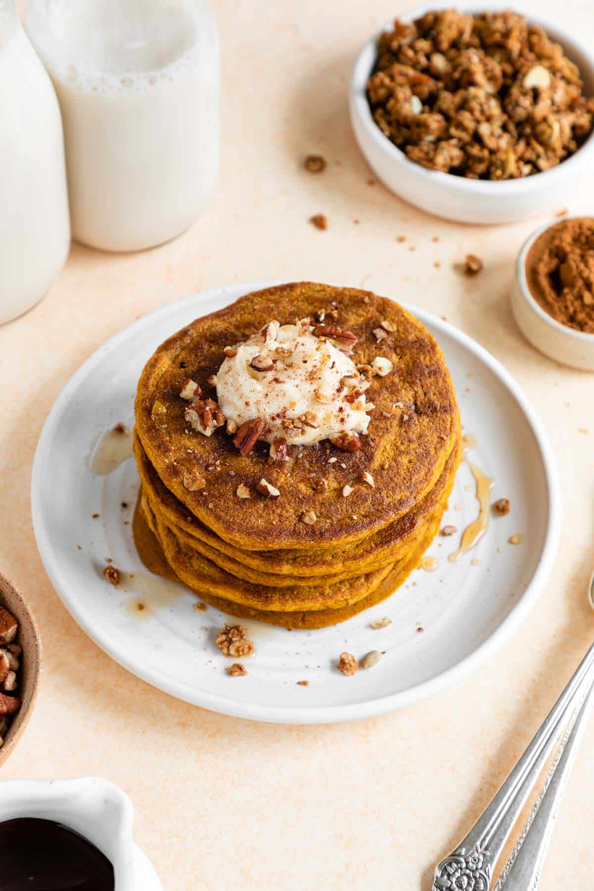 vegan pumpkin pancakes on a plate with whipped cream, maple syrup, and pecans on top