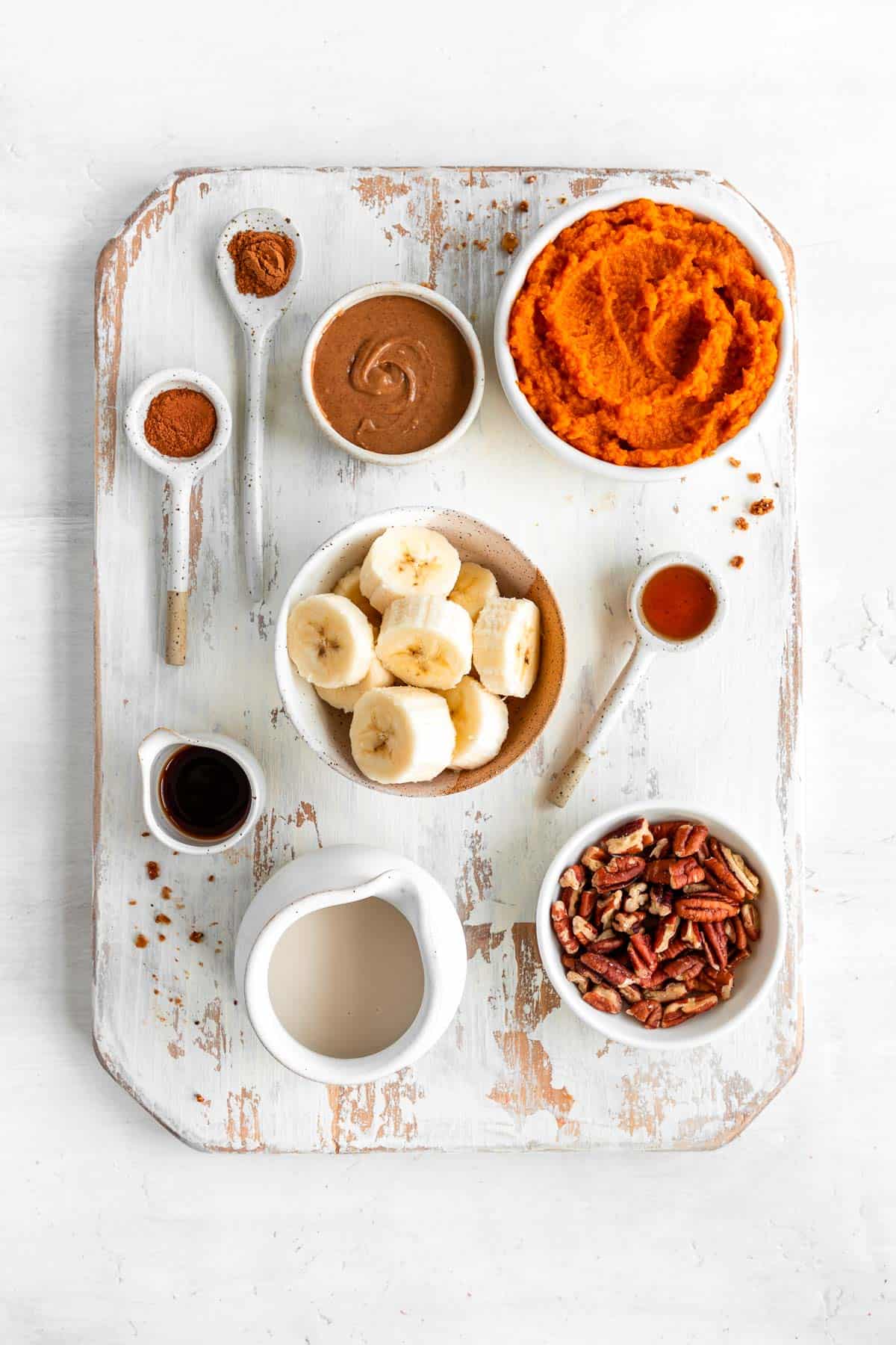 a distressed wood cutting board topped with bowls of pumpkin puree, banana, pecans, oat milk, almond butter, cinnamon, maple syrup, and vanilla