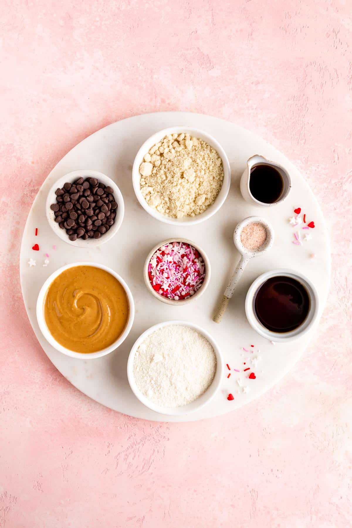 a round white marble slab topped with bowls of cashew butter, almond flour, coconut flour, maple syrup, chocolate chips, and valentine's day sprinkles 