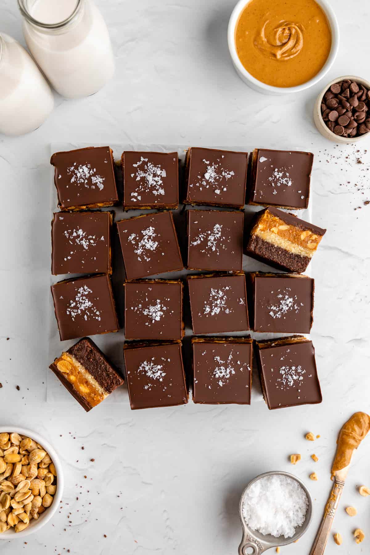 a pile of no bake snickers brownies surrounded by peanut butter, peanuts, and chocolate chips