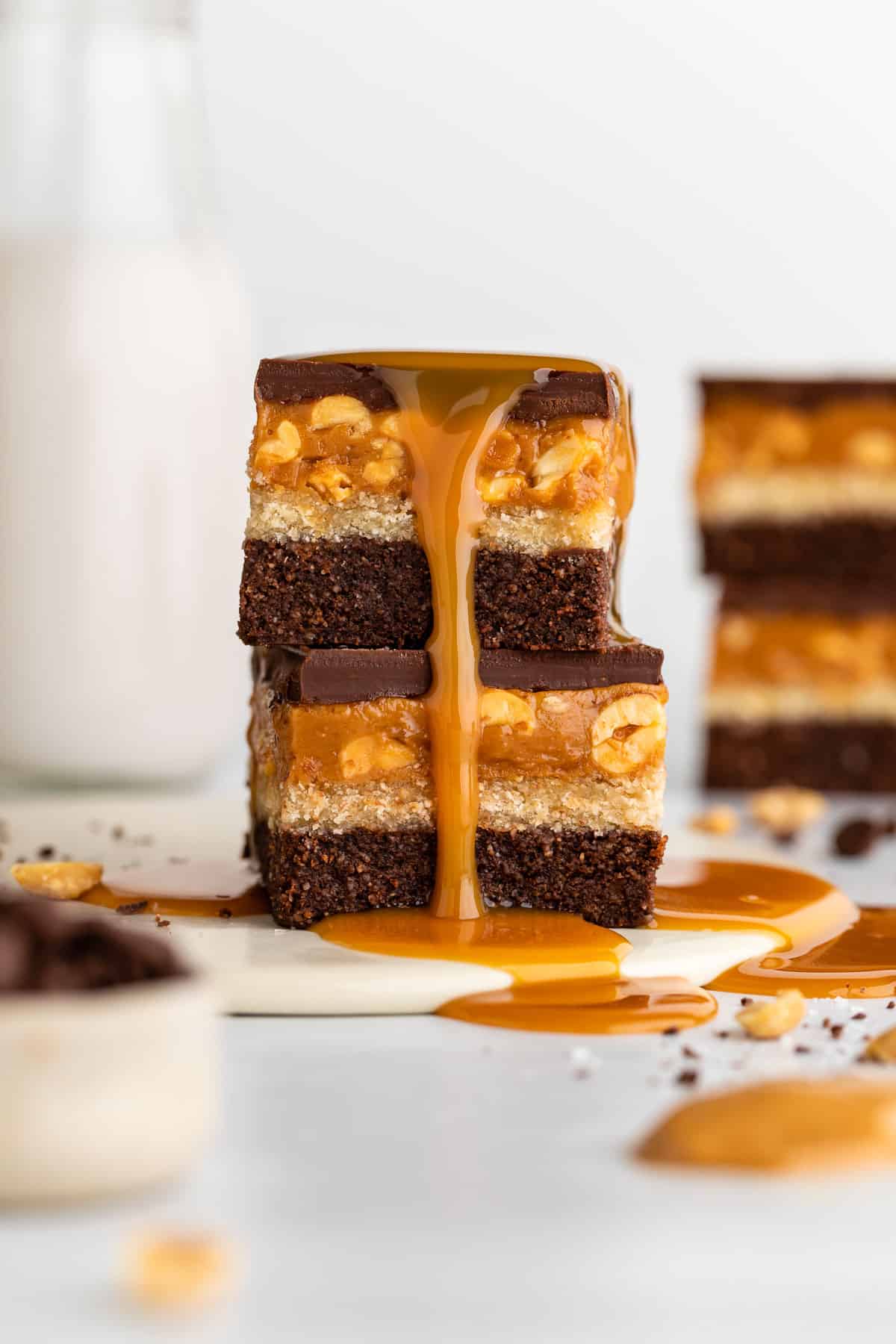 a stack of two vegan no bake snickers brownies with caramel poured over them