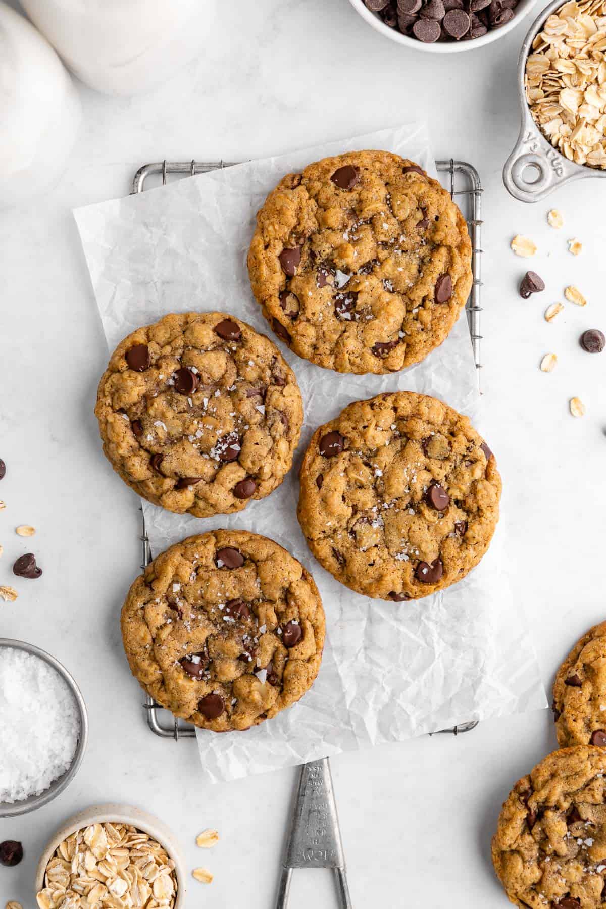 four vegan oatmeal chocolate chip cookies on a wire cooling rack surrounded by oats, chocolate chips, and flaky sea salt