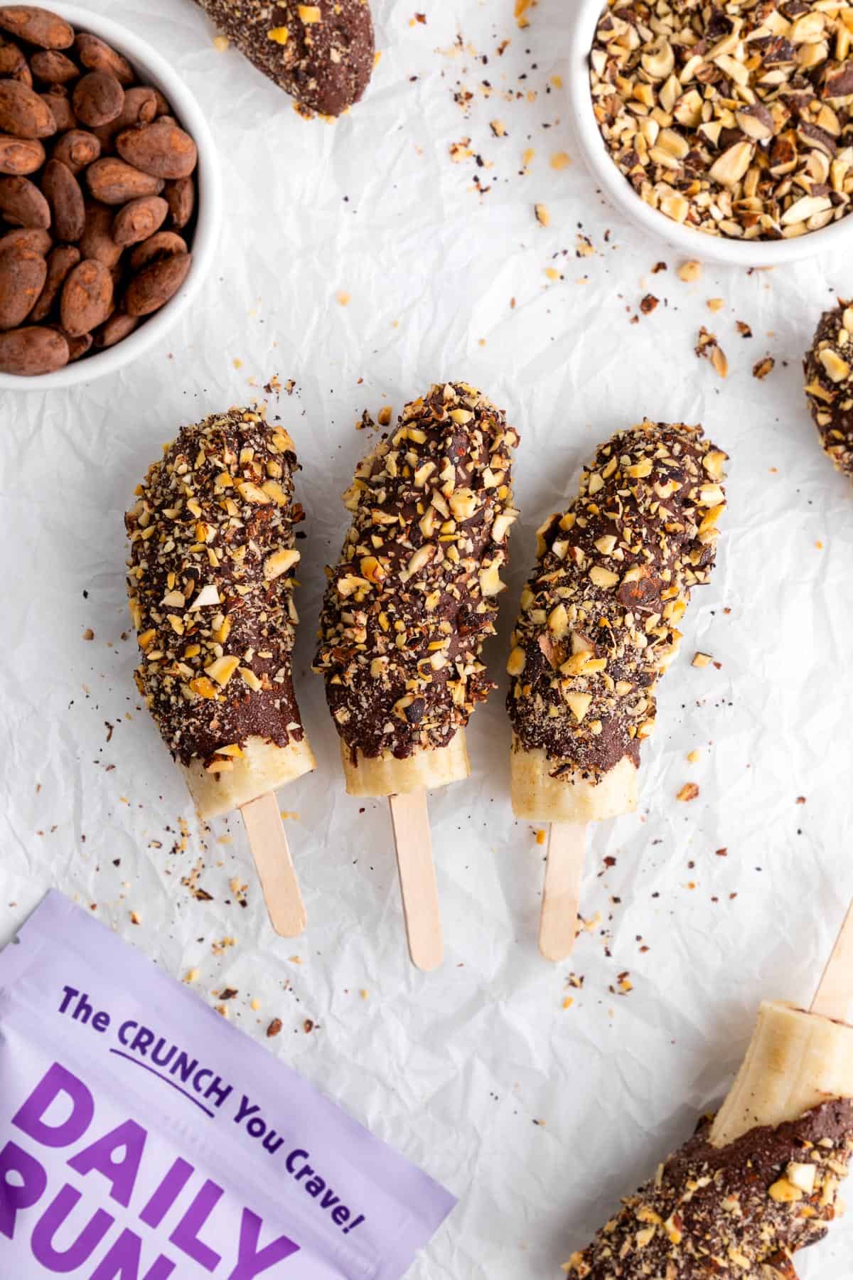 three chocolate covered frozen bananas on popsicle sticks coated in crushed nuts