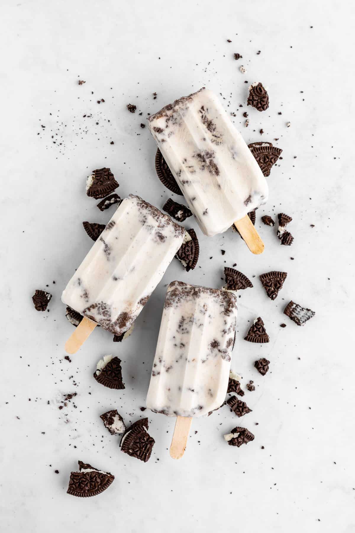 three vegan cookies and cream popsicles laying on top of crushed Oreos