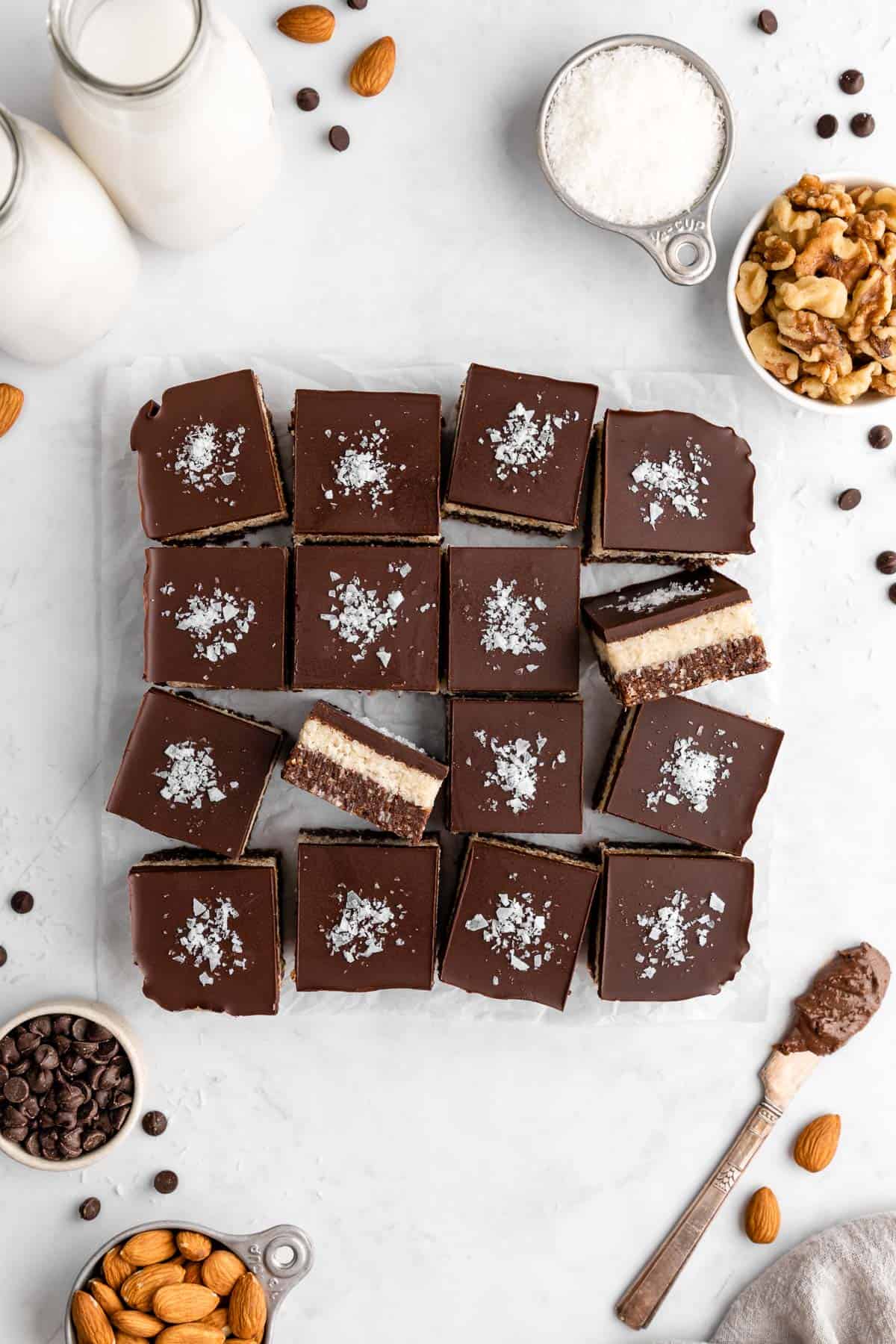 a pile of no-bake coconut brownies surrounded by chocolate chips, almonds, and walnuts