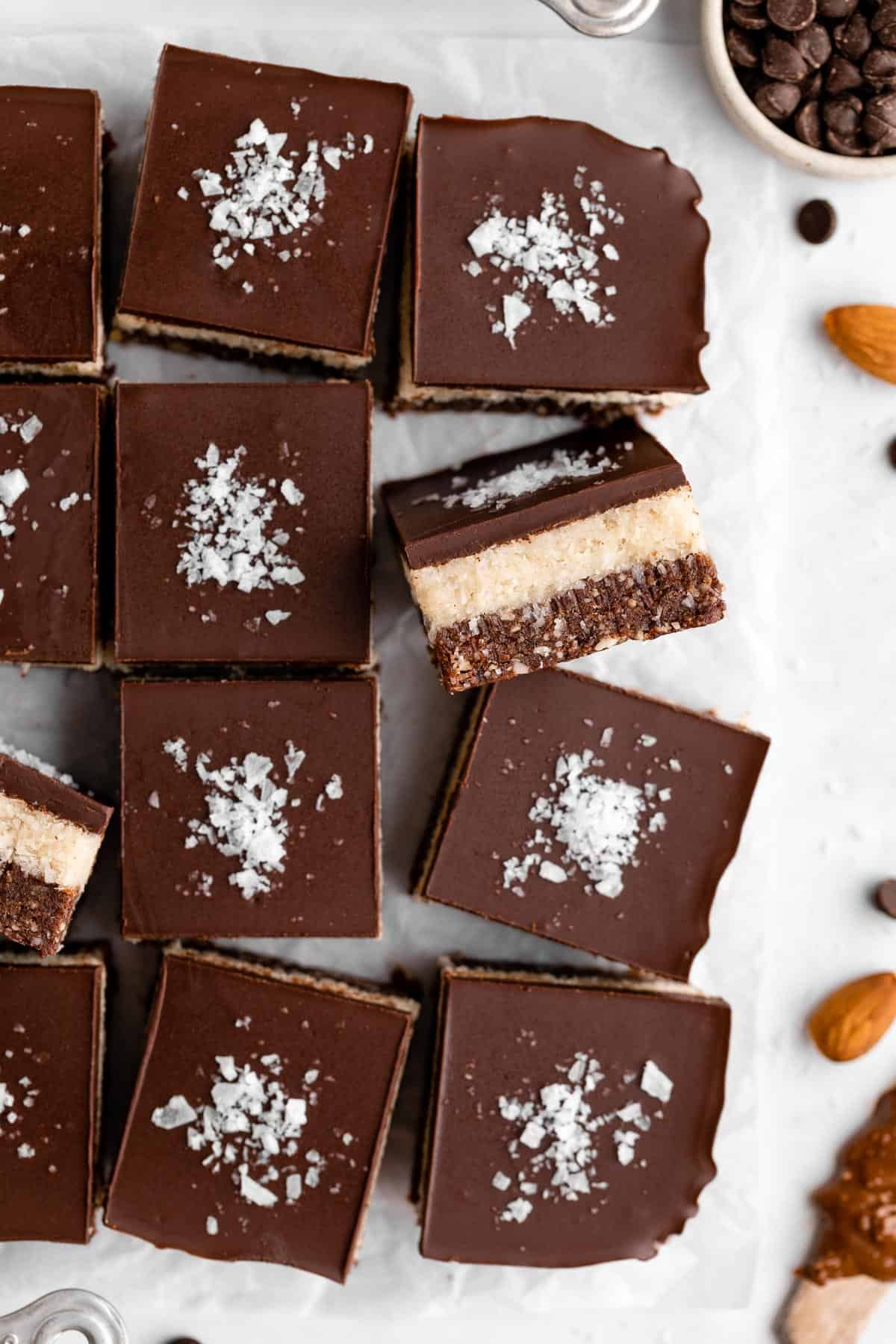 a pile of layered no-bake coconut brownies surrounded by almonds, walnuts, and chocolate chips