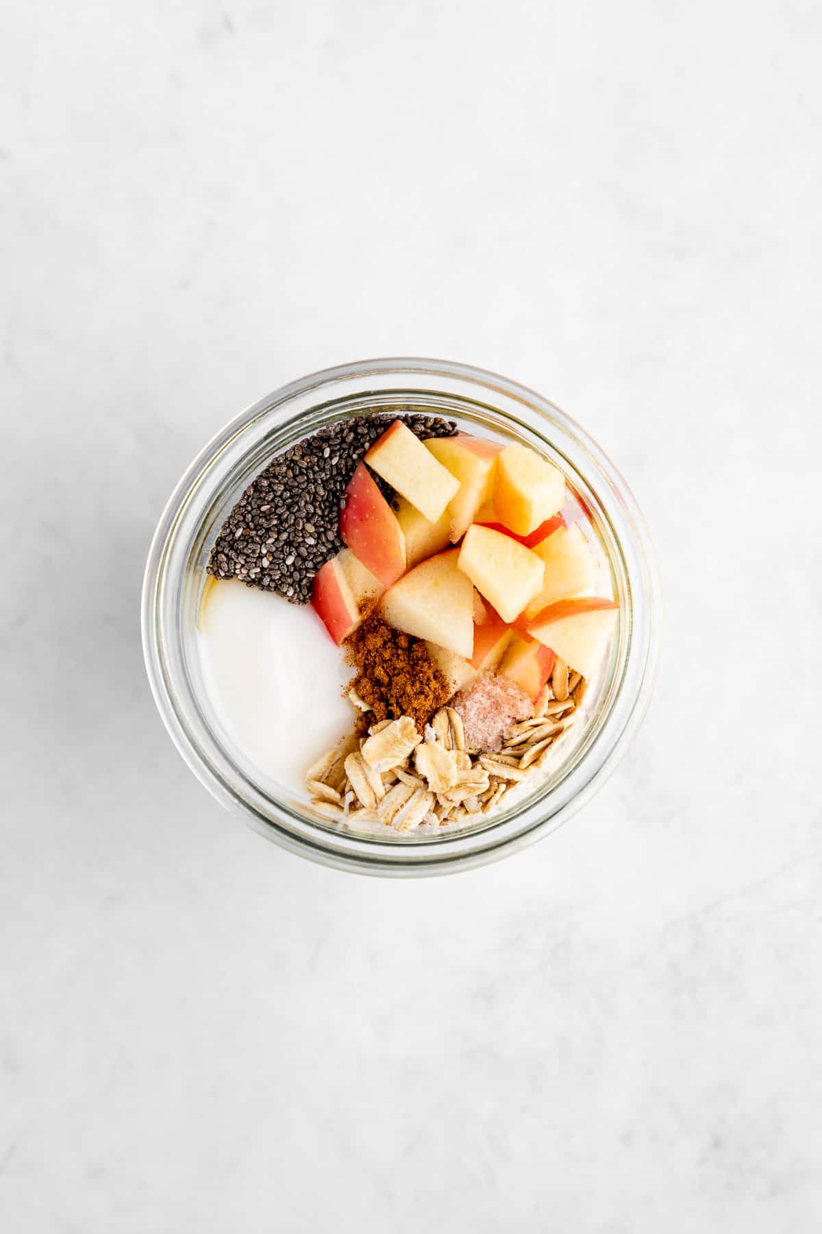 overhead photo of a mason jar filled with rolled oats, diced apples, yogurt, chia seeds, cinnamon, and maple syrup