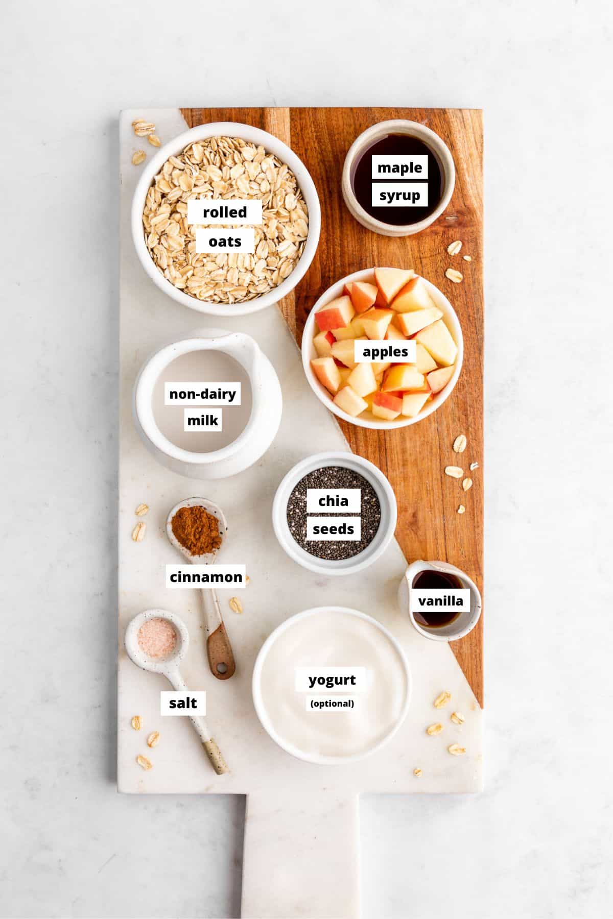 a marble serving board topped with bowls of oats, apples, maple syrup, yogurt, chia seeds, and cinnamon