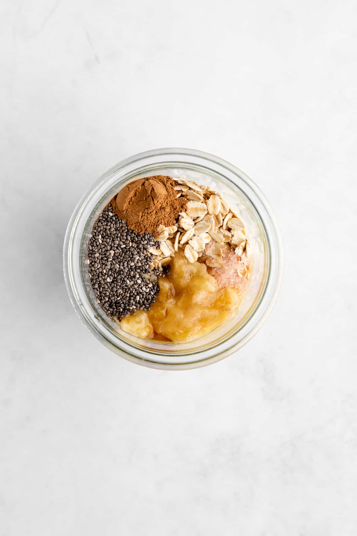 overhead photo of a mason jar filled with rolled oats, mashed banana, almond milk, chia seeds, cinnamon, and maple syrup