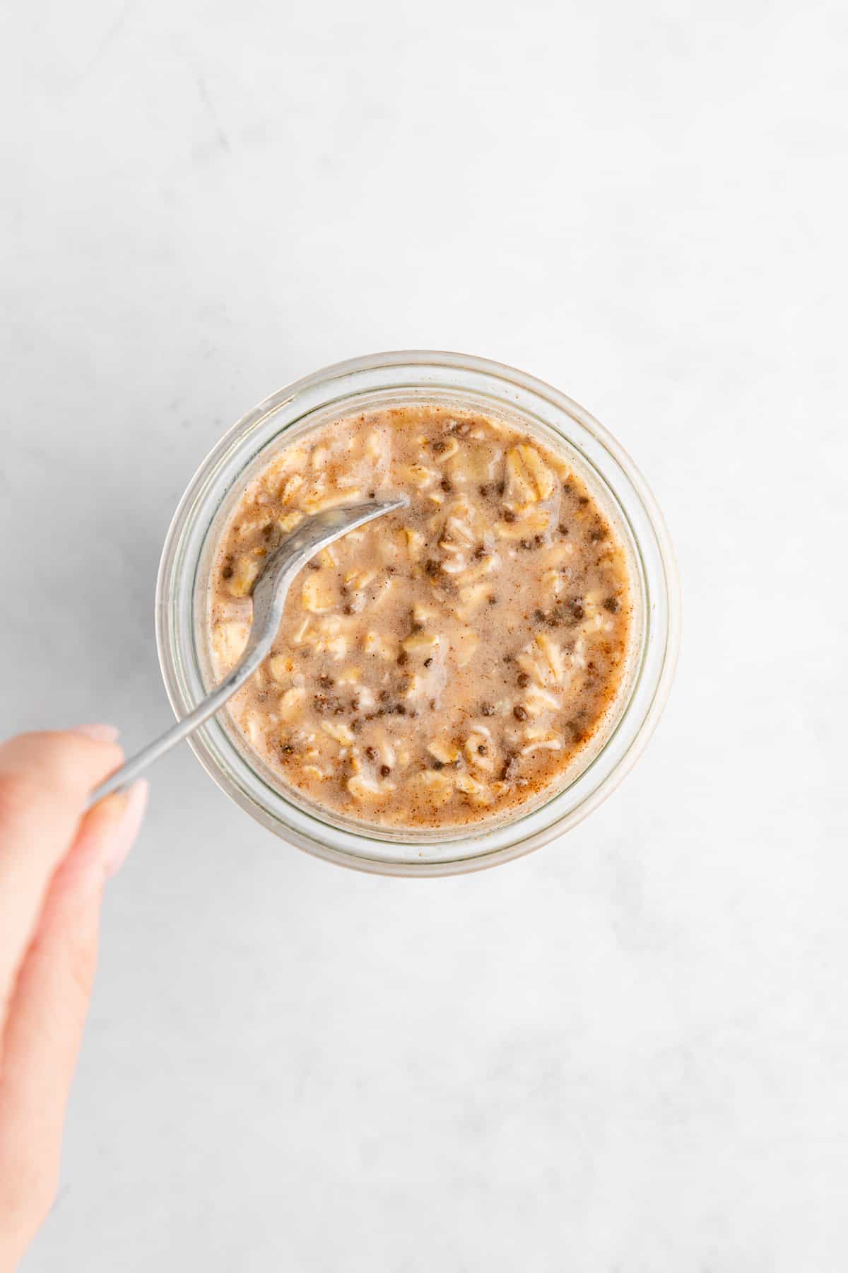 a left hand using a spoon to mix banana overnight oats in a jar