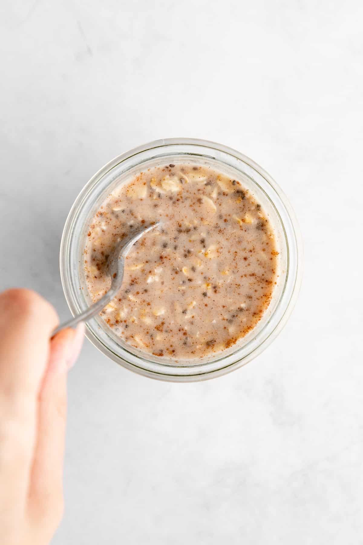 left hand holding a spoon mixing basic overnight oats in a jar