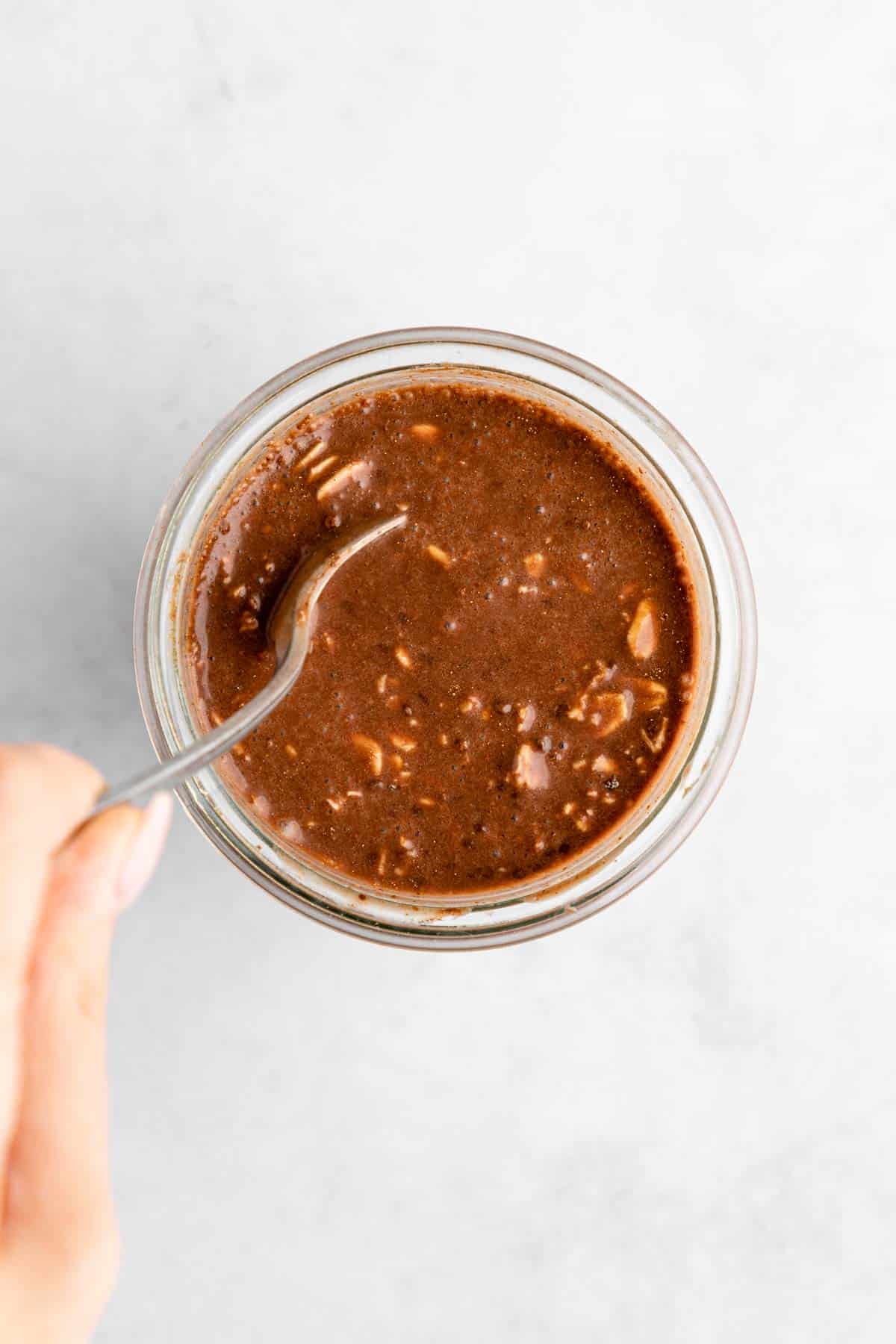left hand holding a spoon mixing vegan chocolate overnight oats in a mason jar