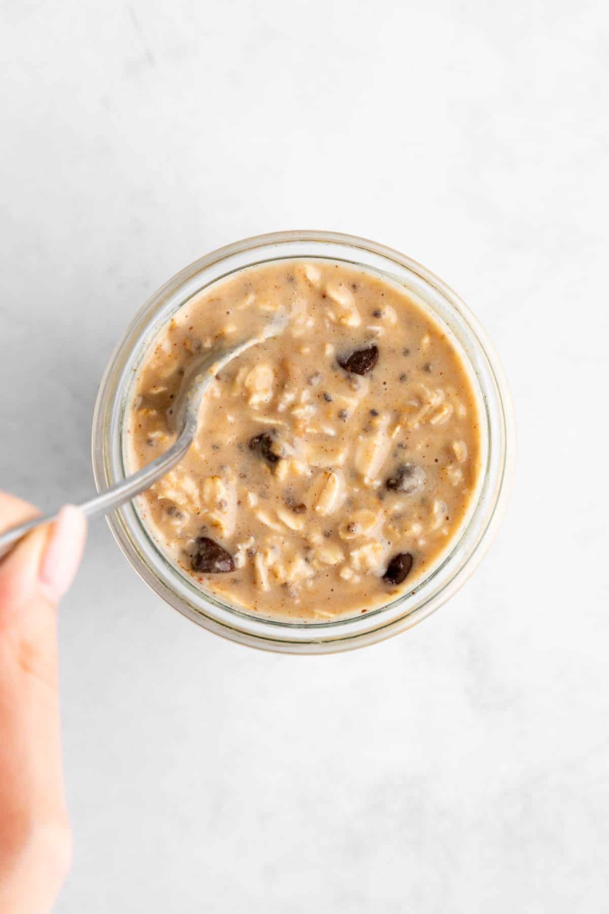 a left hand holding a spoon mixing vegan cookie dough overnight oats in a jar